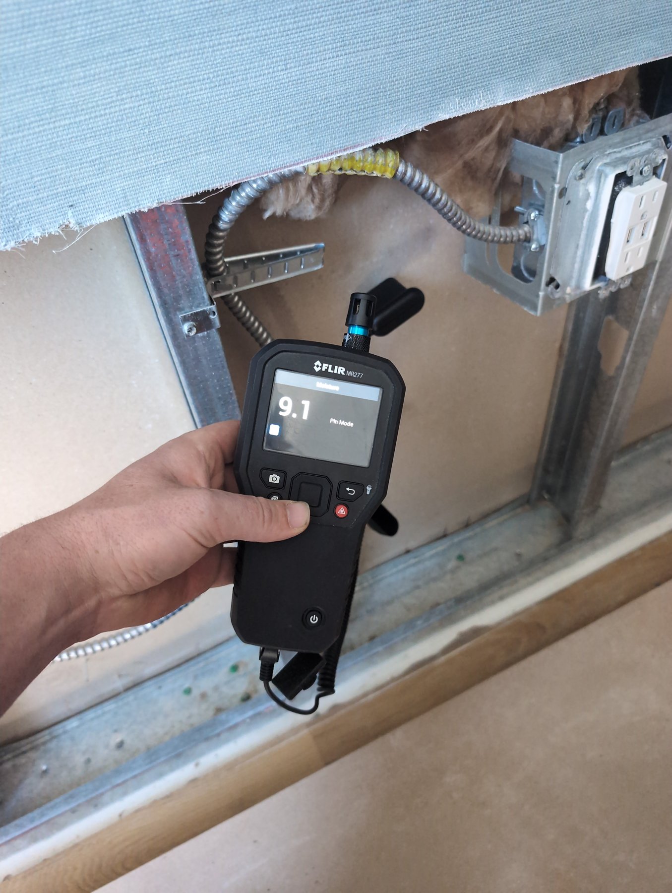 Inspector reading a FLIR pin moisture meter on an opened wall cavity during a water damage inspection
